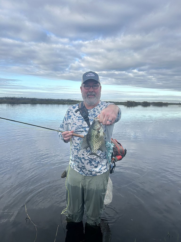 A man in fishing gear holding a speck fish he caught in a peaceful river setting.