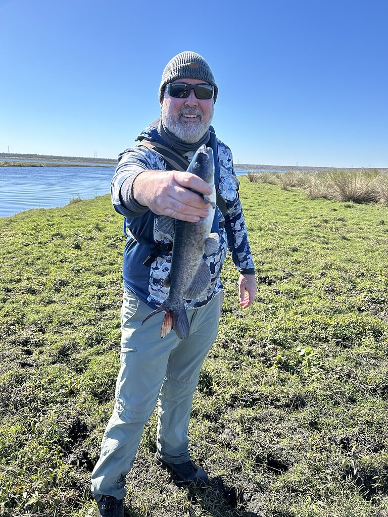 Man holding a freshly caught catfish by the riverbank.