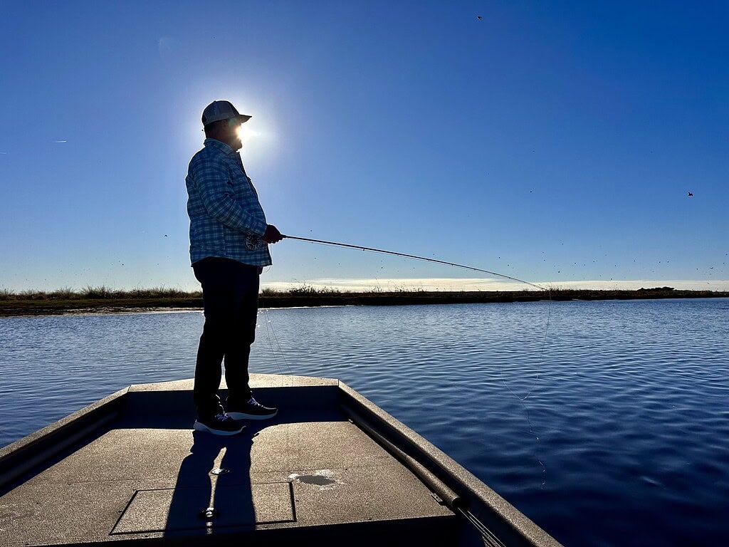 A fisherman stands on a boat casting his line on a calm river during sunset, enjoying fly fishing for American and Hickory Shad.